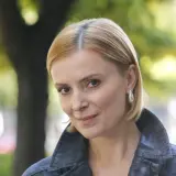 Uśmiechnięta Halina Mlynkova, której wiek dodaje jej uroku, w jeansowej kurtce na tle zieleni.