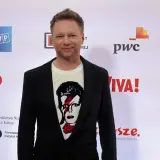 Maciej Stuhr, znany z wielu ról, prezentuje się na ściance. Jego wzrost jest imponujący, a koszulka z Davidem Bowiem dodaje mu charakteru.