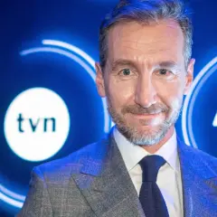 Uśmiechnięty mężczyzna w garniturze na tle logo TVN. Jeden z dziennikarzy Wiadomości TVN, gotowy do przekazania najnowszych informacji.