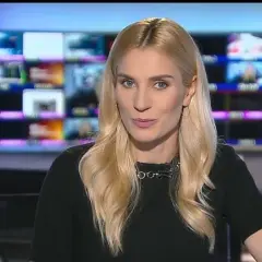 Blond dziennikarka TVN w czarnej bluzce z naszyjnikiem, na tle monitorów.
