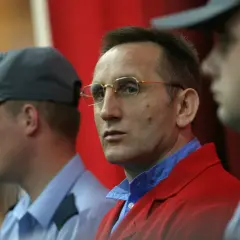 Słowik gangster w czerwonej marynarce, otoczony przez ochroniarzy w niebieskich koszulach i czapkach.