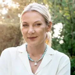 Uśmiechnięta Martyna Kupczyk w białej marynarce i naszyjniku z pereł. W tle zielony ogród.