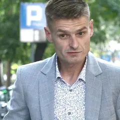 Tomasz Komenda w szarym garniturze i wzorzystej koszuli, z widocznym znakiem P. Jego wiek jest trudny do określenia na pierwszy rzut oka.