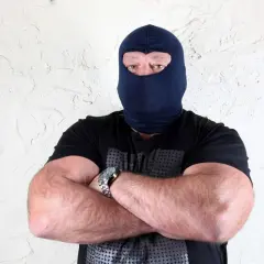 Masywny mężczyzna w kominiarce, z rękami skrzyżowanymi na piersi, wygląda jak groźny gangster.