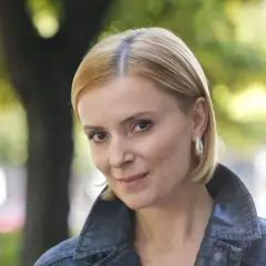 Uśmiechnięta Halina Mlynkova, której wiek dodaje jej uroku, w jeansowej kurtce na tle zieleni.
