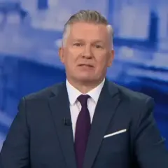 Jeden z prezenterów Fakty TVN w granatowym garniturze i bordowym krawacie, na tle niebieskiej grafiki.