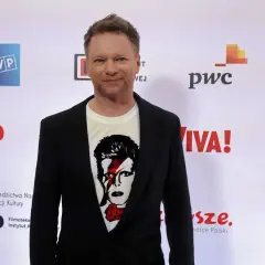Maciej Stuhr, znany z wielu ról, prezentuje się na ściance. Jego wzrost jest imponujący, a koszulka z Davidem Bowiem dodaje mu charakteru.