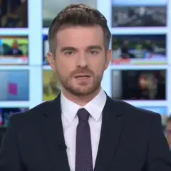Łukasz Jedliński, dziennikarz, w garniturze i krawacie, na tle ekranów informacyjnych. Jego życiorys to historia kariery w mediach.