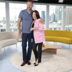 Ewa Drzyzga z mężem w studiu telewizyjnym, uśmiechnięci, na tle okien z widokiem na miasto.