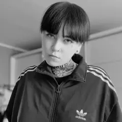 Czarno-białe zdjęcie Patrycji Widera w bluzie z kapturem Adidas. Jej twarz zdobią tatuaże i kolczyki.