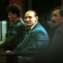 Trzech mężczyzn w garniturach siedzi w sali sądowej. Jeden z nich, w okularach, wygląda jak pershing gangster.