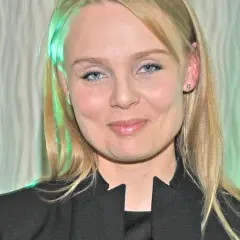 Patrycja Redo, z niebieskimi oczami i blond włosami, uśmiecha się. W tle falista zasłona.