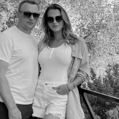 Aryna Sabalenka z partnerem, uśmiechnięci, w okularach przeciwsłonecznych, na tle drzew i architektury.