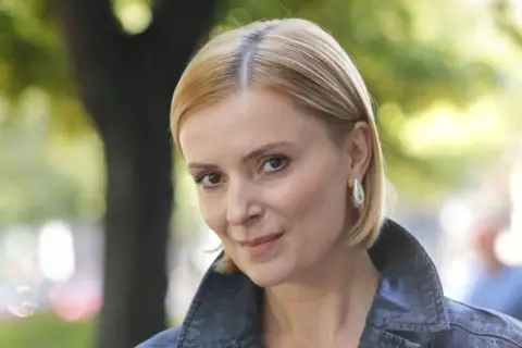 Uśmiechnięta Halina Mlynkova, której wiek dodaje jej uroku, w jeansowej kurtce na tle zieleni.