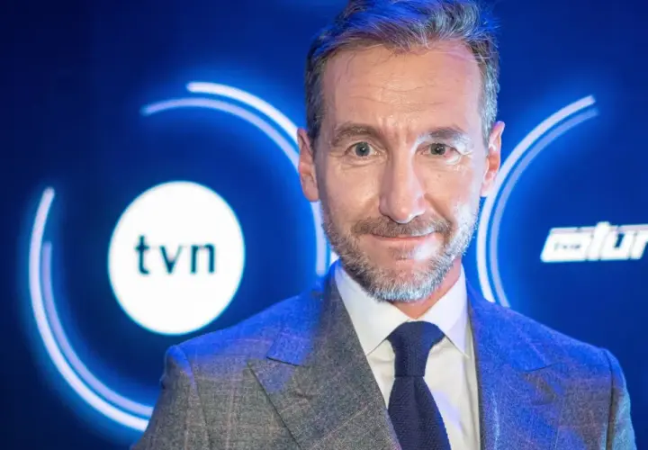 Uśmiechnięty mężczyzna w garniturze na tle logo TVN. Jeden z dziennikarzy Wiadomości TVN, gotowy do przekazania najnowszych informacji.