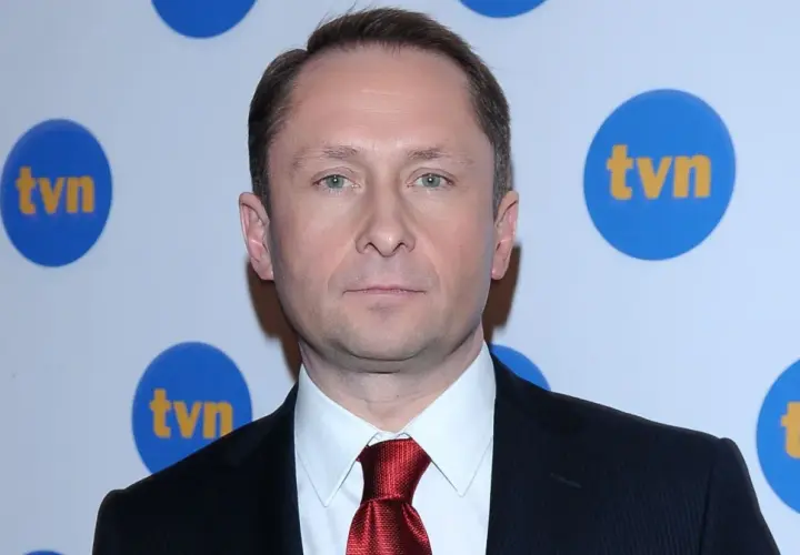 Kamil Durczok w garniturze i czerwonym krawacie na tle logo TVN. Prowadzący Fakty TVN.