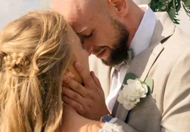 Agnieszka Kaczorowska i jej mąż w czułym uścisku podczas ślubu. Ona ma blond włosy splecione w warkocz, on w jasnej marynarce z butonierką.