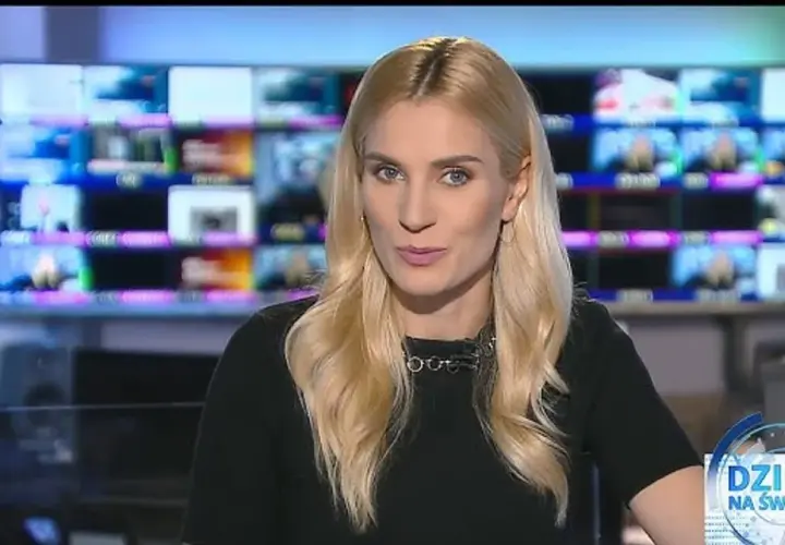 Blond dziennikarka TVN w czarnej bluzce z naszyjnikiem, na tle monitorów.
