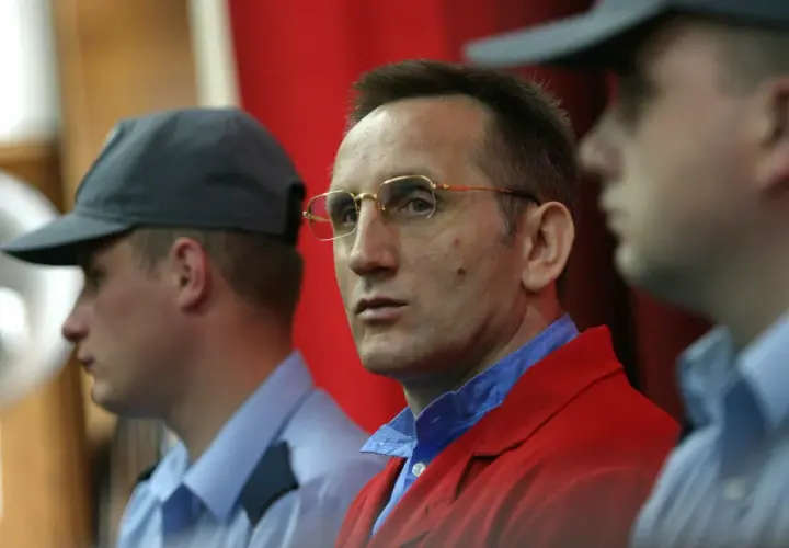 Słowik gangster w czerwonej marynarce, otoczony przez ochroniarzy w niebieskich koszulach i czapkach.
