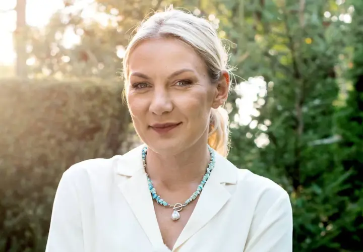 Uśmiechnięta Martyna Kupczyk w białej marynarce i naszyjniku z pereł. W tle zielony ogród.