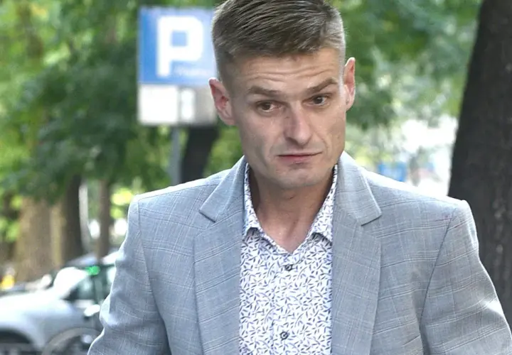 Tomasz Komenda w szarym garniturze i wzorzystej koszuli, z widocznym znakiem P. Jego wiek jest trudny do określenia na pierwszy rzut oka.
