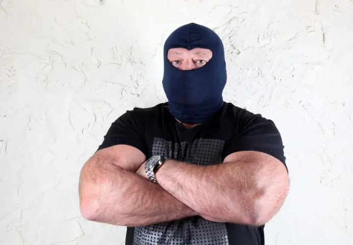 Masywny mężczyzna w kominiarce, z rękami skrzyżowanymi na piersi, wygląda jak groźny gangster.