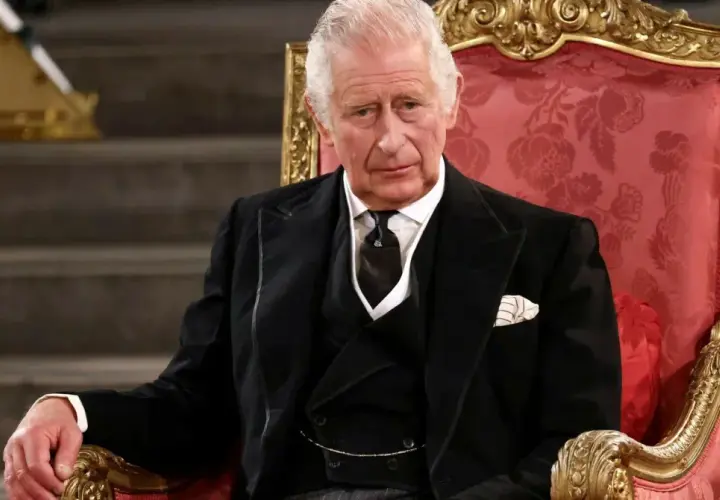 Król Karol w wieku siedemdziesięciu lat, w eleganckim czarnym garniturze, siedzi na ozdobnym tronie.