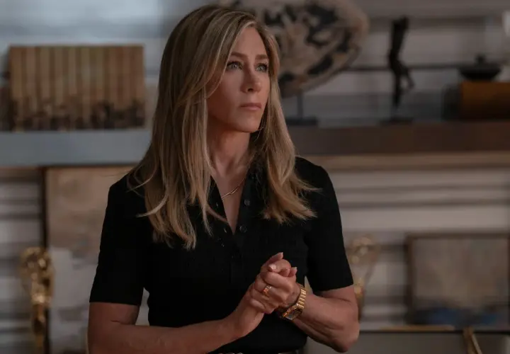 Jennifer Aniston zamyślona, w czarnej bluzce, z dłońmi złożonymi na piersi. W tle widać elementy wystroju wnętrza, może nawiązujące do jej życia, jakby myślała o swoich przyszłych dzieciach.