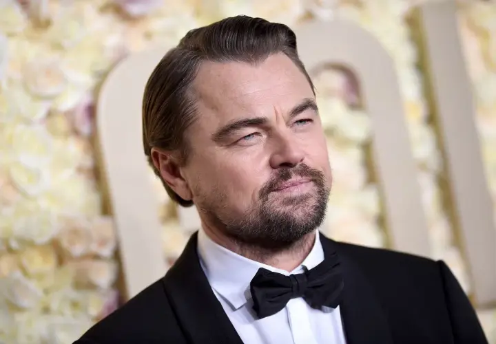 Leonardo DiCaprio wiek: aktor w czarnym smokingu z muszką, z siwiejącymi włosami i zarostem, na tle kwiatowej dekoracji.