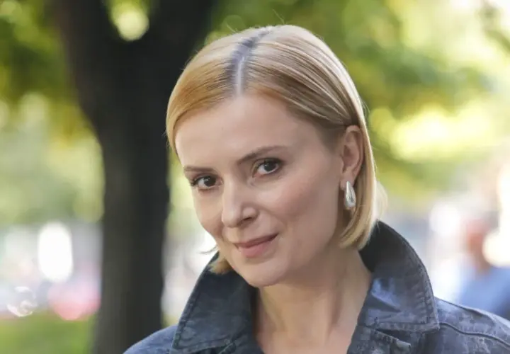 Uśmiechnięta Halina Mlynkova, której wiek dodaje jej uroku, w jeansowej kurtce na tle zieleni.