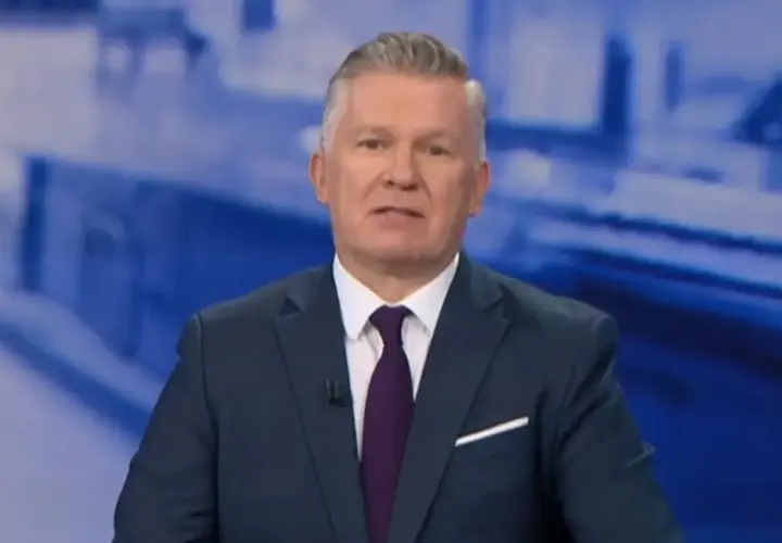 Jeden z prezenterów Fakty TVN w granatowym garniturze i bordowym krawacie, na tle niebieskiej grafiki.