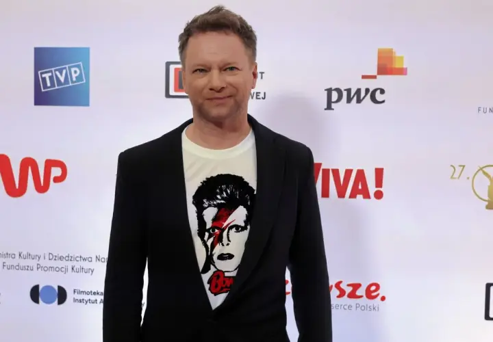Maciej Stuhr, znany z wielu ról, prezentuje się na ściance. Jego wzrost jest imponujący, a koszulka z Davidem Bowiem dodaje mu charakteru.