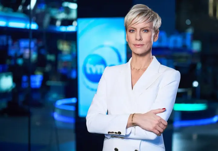 Kobieta w białej marynarce, prezenterka TVN Fakty, stoi na tle studia. Jej spojrzenie jest pewne, a postawa świadczy o profesjonalizmie.