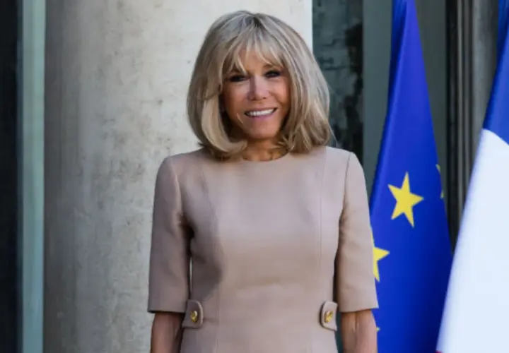 Brigitte Macron z uśmiechem i charakterystycznymi blond włosami, na tle flag UE i Francji.
