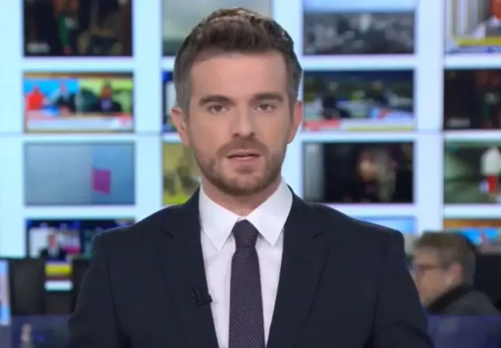 Łukasz Jedliński, dziennikarz, w garniturze i krawacie, na tle ekranów informacyjnych. Jego życiorys to historia kariery w mediach.