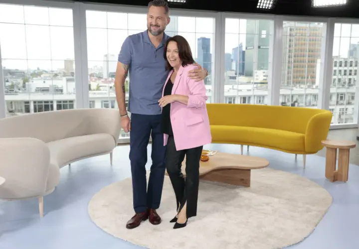Ewa Drzyzga z mężem w studiu telewizyjnym, uśmiechnięci, na tle okien z widokiem na miasto.
