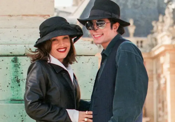 Lisa Marie Presley i jej mąż Michael Jackson w kapeluszach i okularach przeciwsłonecznych, uśmiechnięci na tle budowli.