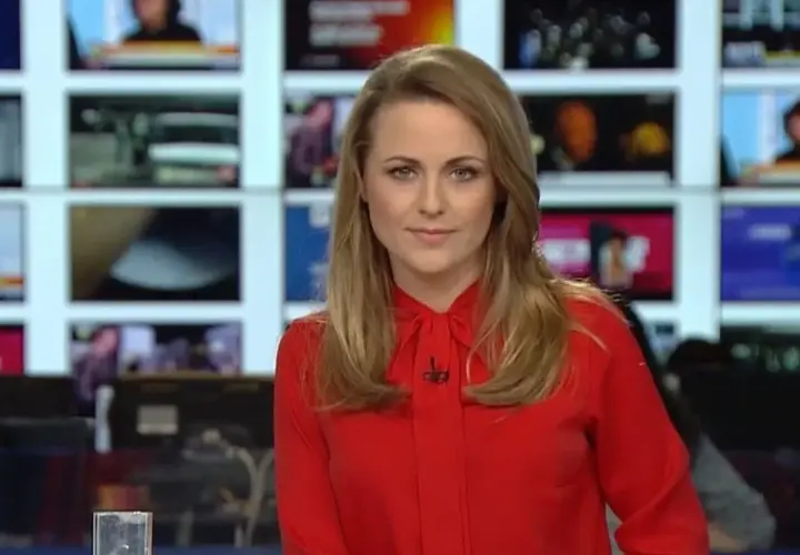 Marta Kłos, w czerwonej bluzce, na tle ekranów informacyjnych. Jej biografia to historia kariery w mediach.