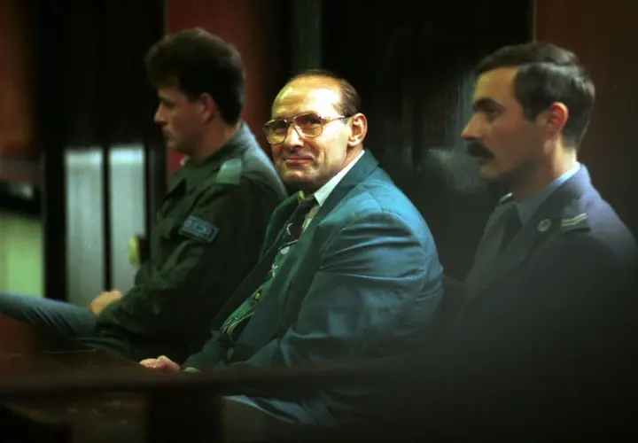 Trzech mężczyzn w garniturach siedzi w sali sądowej. Jeden z nich, w okularach, wygląda jak pershing gangster.
