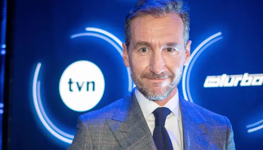 Uśmiechnięty mężczyzna w garniturze na tle logo TVN. Jeden z dziennikarzy Wiadomości TVN, gotowy do przekazania najnowszych informacji.
