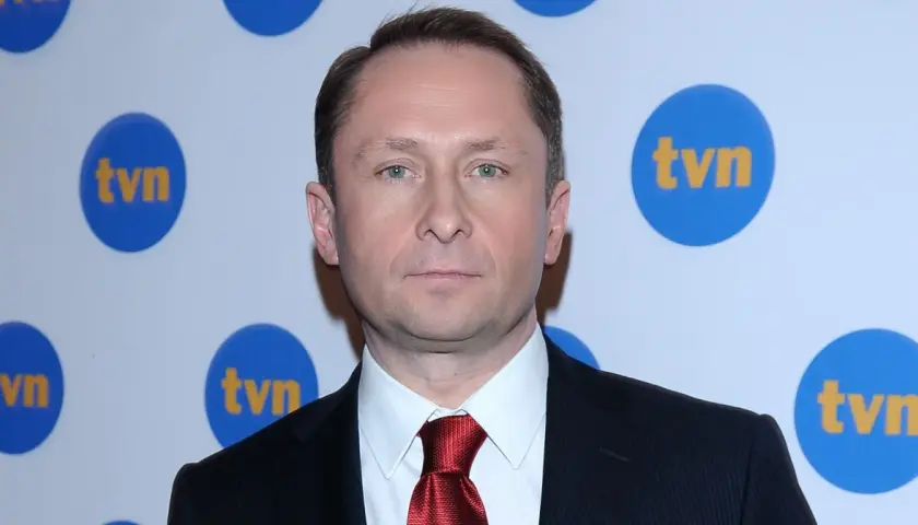 Kamil Durczok w garniturze i czerwonym krawacie na tle logo TVN. Prowadzący Fakty TVN.