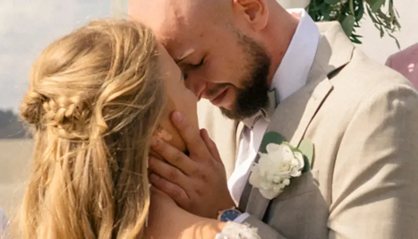 Agnieszka Kaczorowska i jej mąż w czułym uścisku podczas ślubu. Ona ma blond włosy splecione w warkocz, on w jasnej marynarce z butonierką.