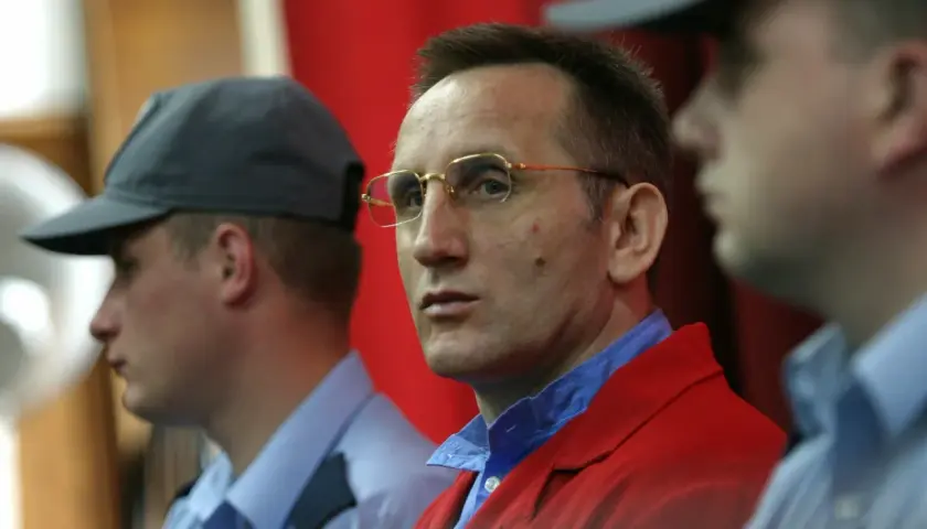 Słowik gangster w czerwonej marynarce, otoczony przez ochroniarzy w niebieskich koszulach i czapkach.