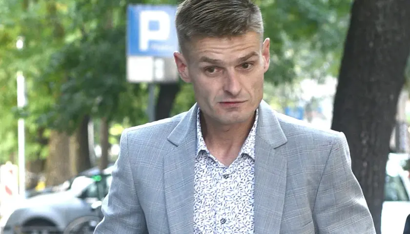 Tomasz Komenda w szarym garniturze i wzorzystej koszuli, z widocznym znakiem P. Jego wiek jest trudny do określenia na pierwszy rzut oka.