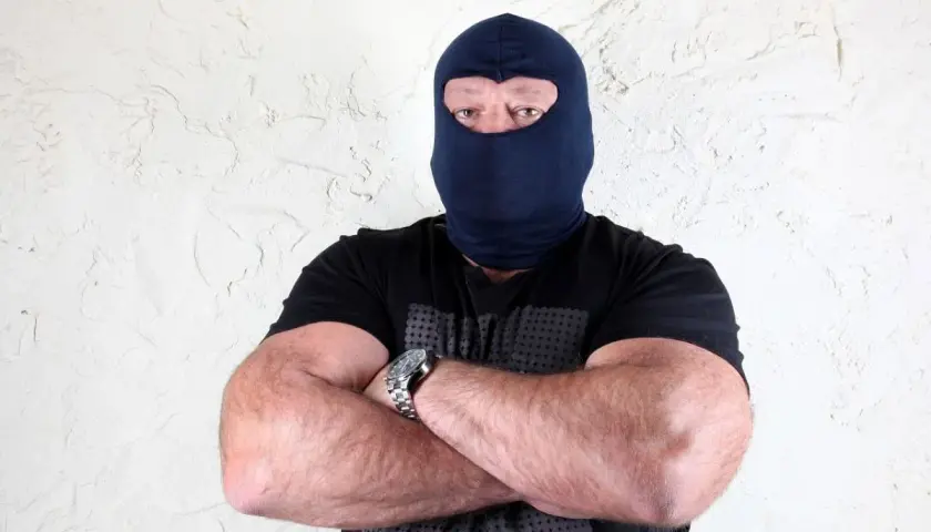 Masywny mężczyzna w kominiarce, z rękami skrzyżowanymi na piersi, wygląda jak groźny gangster.