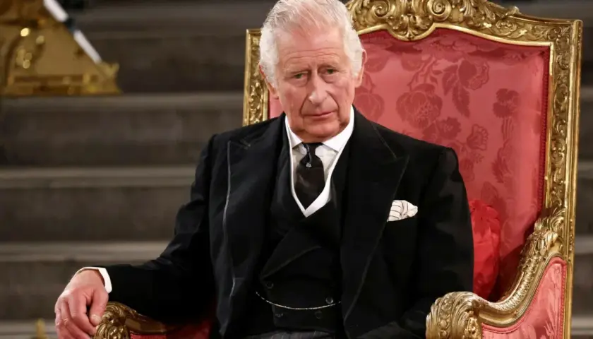 Król Karol w wieku siedemdziesięciu lat, w eleganckim czarnym garniturze, siedzi na ozdobnym tronie.