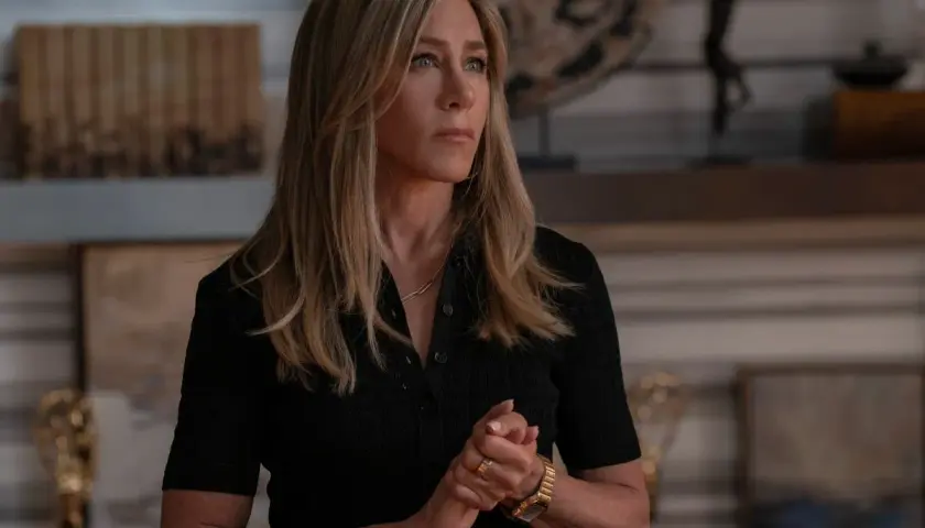 Jennifer Aniston zamyślona, w czarnej bluzce, z dłońmi złożonymi na piersi. W tle widać elementy wystroju wnętrza, może nawiązujące do jej życia, jakby myślała o swoich przyszłych dzieciach.
