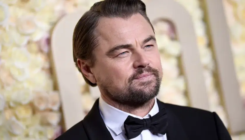 Leonardo DiCaprio wiek: aktor w czarnym smokingu z muszką, z siwiejącymi włosami i zarostem, na tle kwiatowej dekoracji.