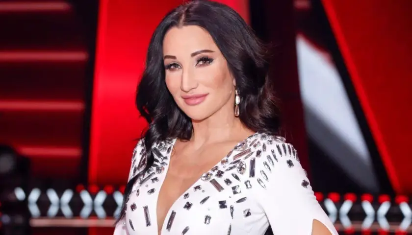 Justyna Steczkowska, której wiek jest tajemnicą, w białej, zdobionej sukni na tle czerwonej sceny.