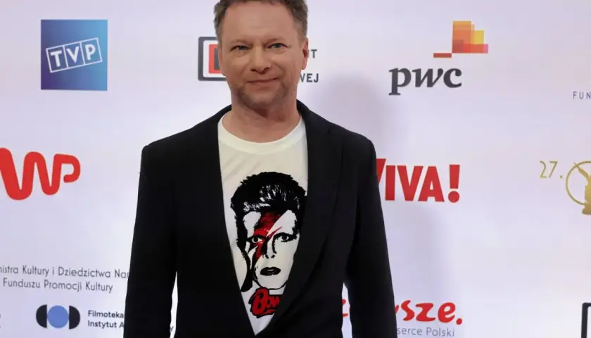 Maciej Stuhr, znany z wielu ról, prezentuje się na ściance. Jego wzrost jest imponujący, a koszulka z Davidem Bowiem dodaje mu charakteru.